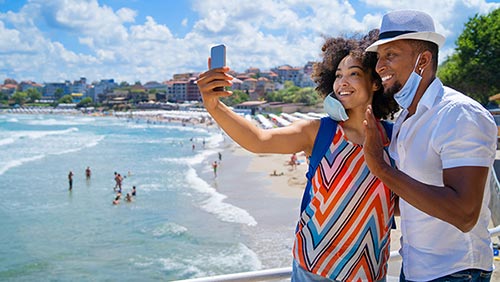 Black couple wearing masks and walking on the beach while taking selfie