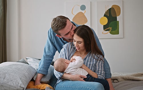 Happy adoptive parents in their bedroom, mother nursing baby with father's support 